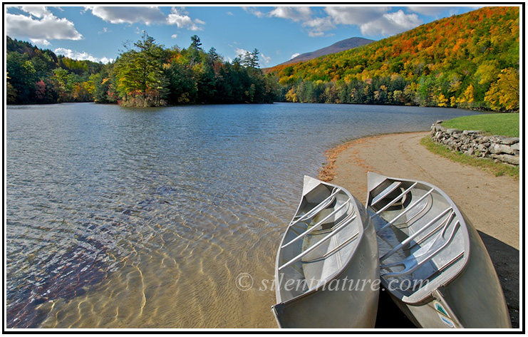 A pair of canoes rest on the shores of a Vermont lake surruounded with autumn colors..