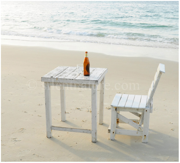 A lone table with a lone chair and a lone bottle on the beach at sunrise.