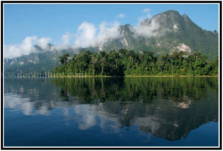 A beautiful karst mountain reflected into the water, fronted by trees and caressed by clouds.