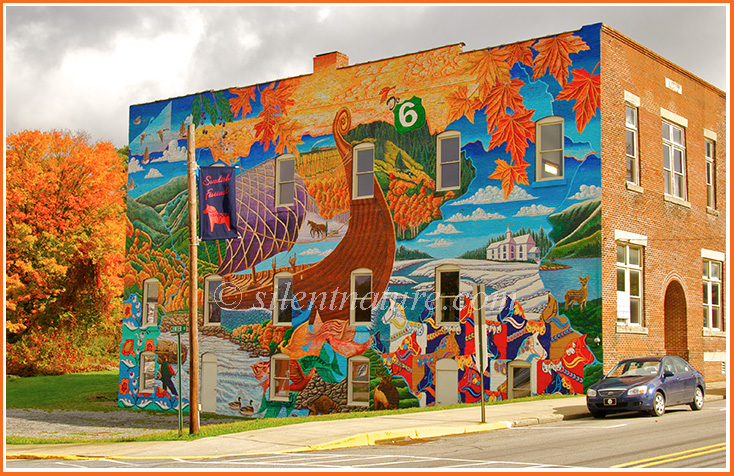 An artist paints a mural on the side of a building the blends the building with the autumn trees.