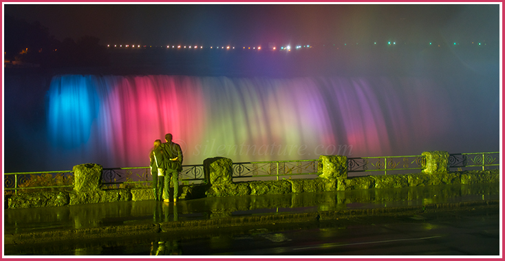 Arm in arm, newly weds overlook Niagra Falls under colred night lights.